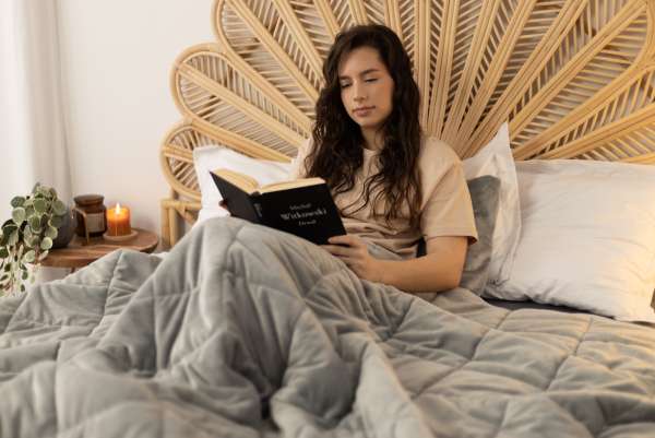 Pige sidder i en seng med et Velourtæppe med tyngde, grå imens hun læser i en bog 