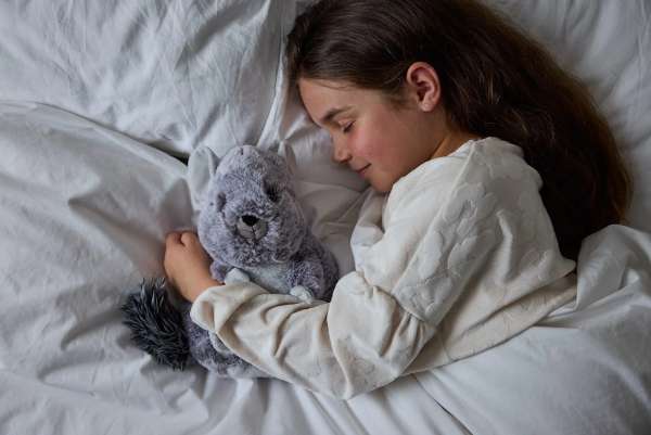 Sansebamse Warmies® Chinchilla ligger i en seng og sover sammen med en pige