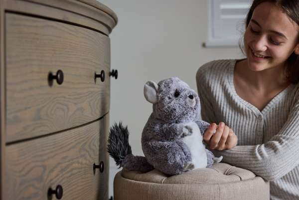 Sansebamse Warmies® Chinchilla sidder på en Puf ved siden af en kommode, og der en pige der holder den i poden