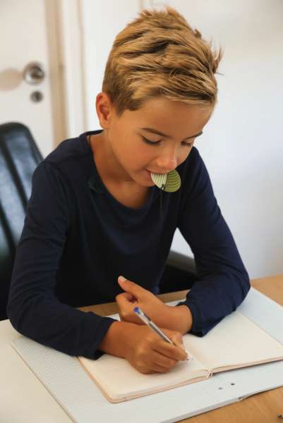 En dreng er i gang med at skrive imens han sidder med et bord samt Han har en CalmCircle 2-i-1 fidget og bideredskab i munden