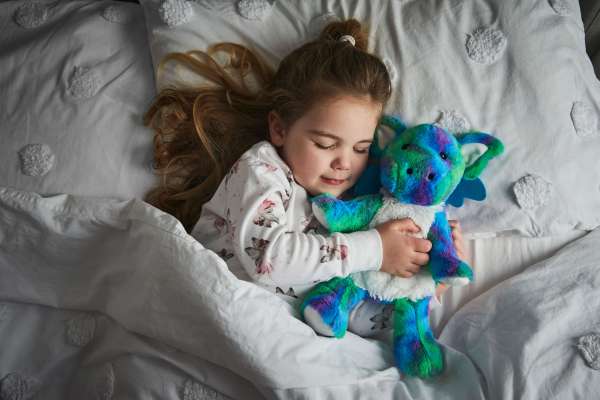 Sansebamse Warmies® regnbuedrage hygge sammen med en pige i en seng